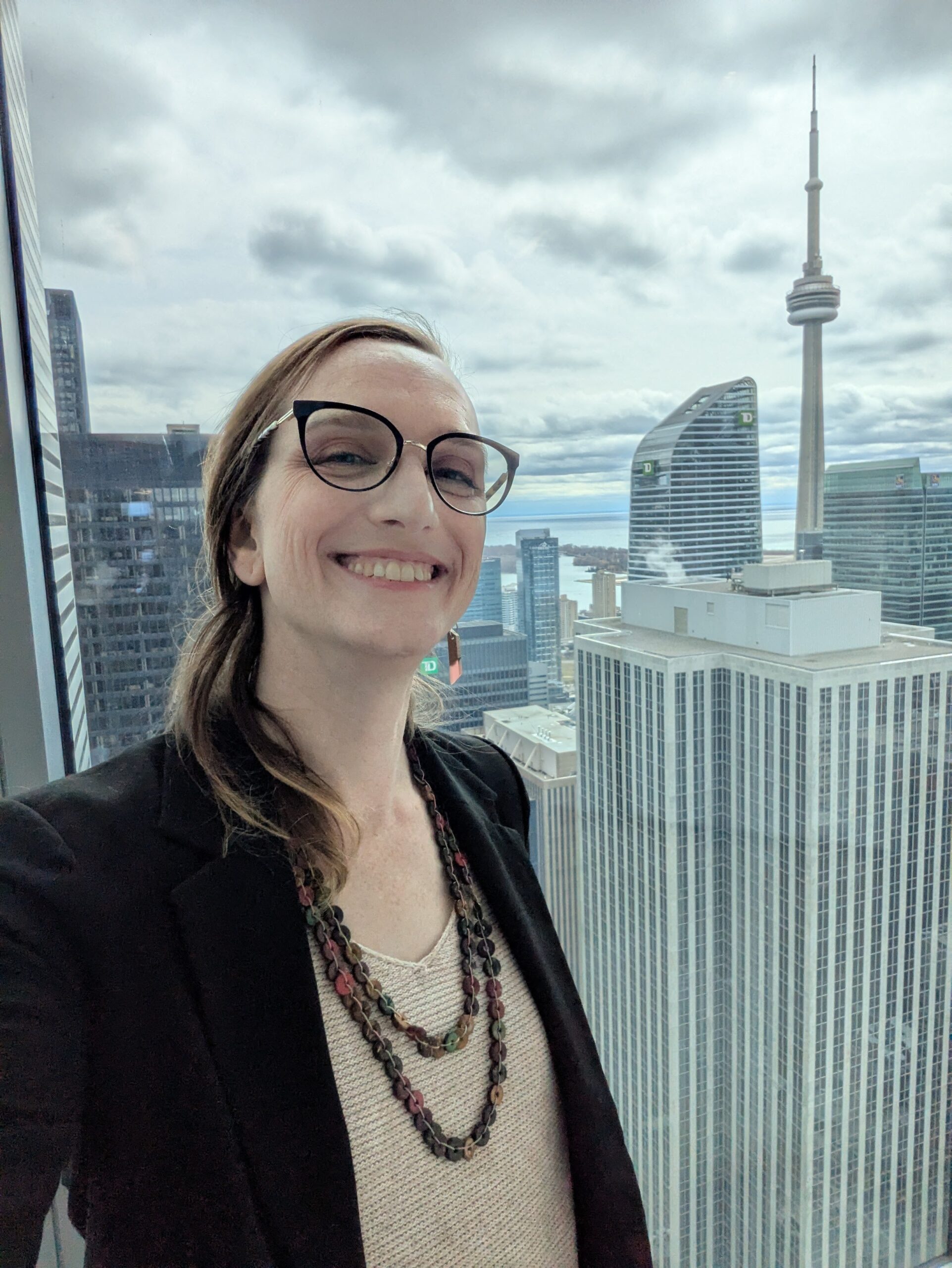 woman with glasses and hair tied back, smiling with view from tall building of CN Tower, Toronto